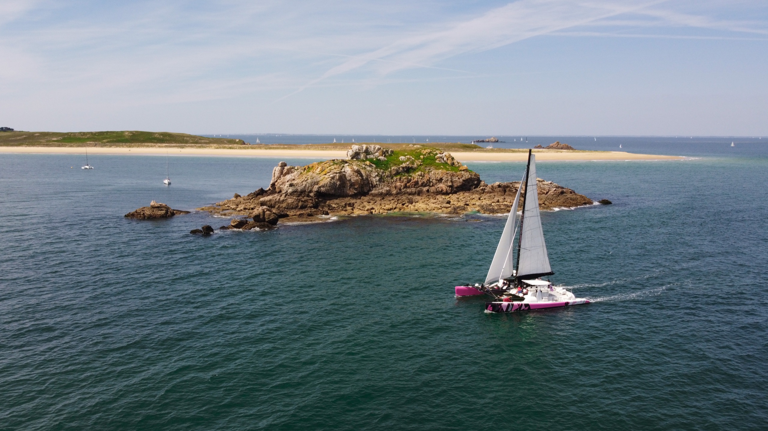 Catamaran longeant un îlot rocheux au large de la Bretagne, vu par drone