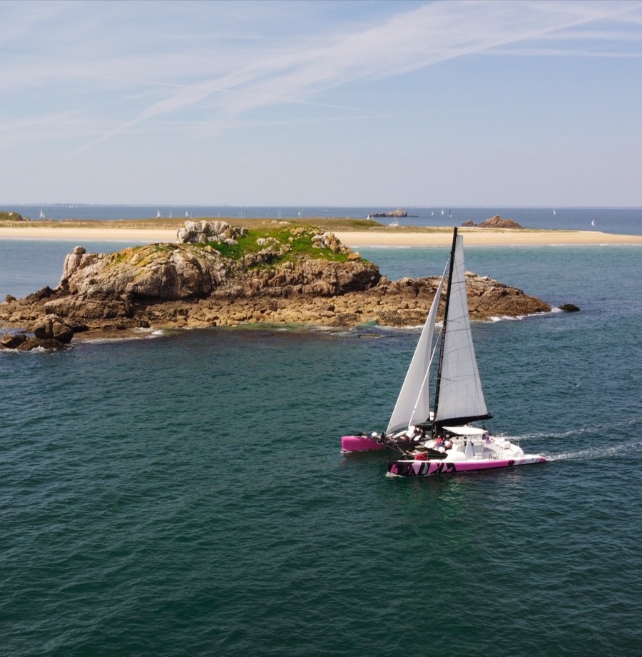Catamaran longeant un îlot rocheux au large de la Bretagne, vu par drone